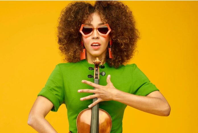 Yilian Cañizares con il violino, abito verde e occhiali da sole su sfondo giallo